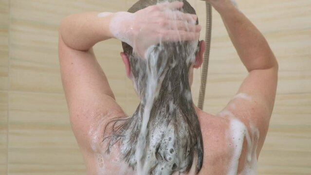 Woman washing her hair, rinsing off foamy shampoo under shower while standing in bathroom, back view. Hair care concept.