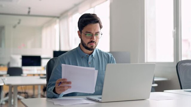 Young busy latin professional business man checking document working at laptop computer in office. Serious businessman accountant expert reading legal paper company file overview at workplace.