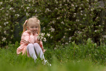 Happy smiling little blonde girl with two ponytails holds blooming flowers and sits on grass. Spring day
