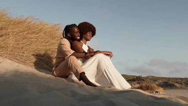 A content couple enjoys a serene moment, embracing on sandy dunes with smiles, portraying love and togetherness.