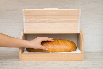 Young adult woman hand holding fresh white bread and taking out from wooden bread box on table top at home kitchen. Closeup. Front view. - obrazy, fototapety, plakaty