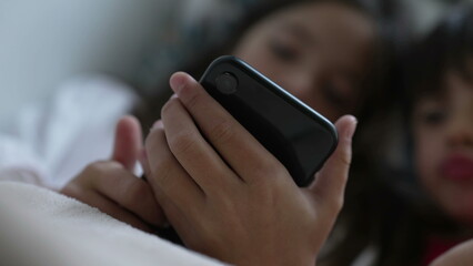 Child close-up hand holding cellphone device absorbed by media entertainment online, blurred kids...