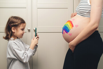 A girl takes a photograph of her pregnant mother. Expecting a child. A happy family. A girl takes a photograph of a drawing on her mother's belly