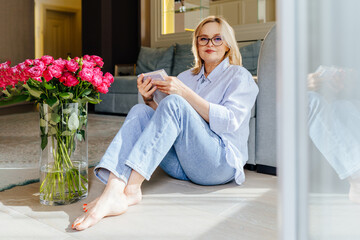 Mature 55 year old woman well-groomed blonde woman with glasses, dressed in jeans and linen blue shirt, sitting on the living room floor, reading a book.