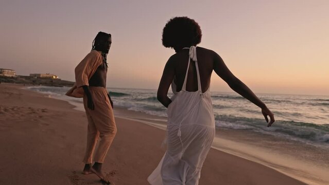 Elegant black couple strolls along a beach with waves at sunset, embodying love, togetherness, and serenity.