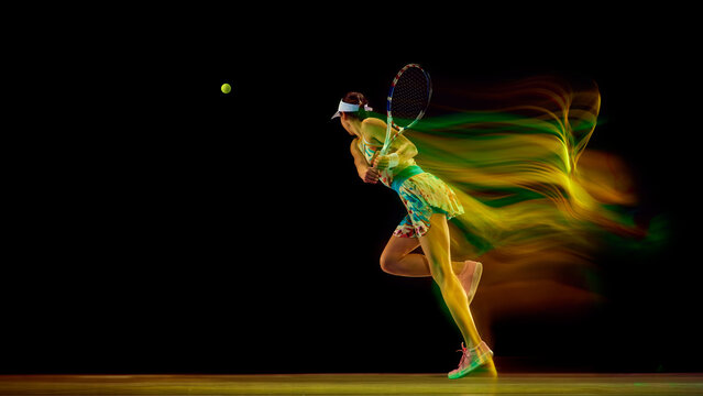 Dynamic photo of woman, tennis player leaping to hit overhead shot in colorful motion blur against black studio background. Concept of sport, active lifestyles, tournaments, energy, movement.