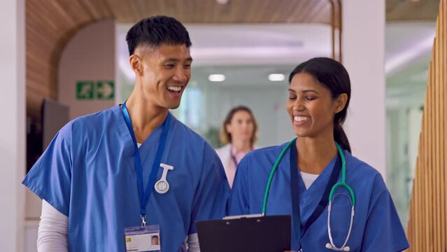 Two Doctors Or Nurses Wearing Scrubs Talking As They Walk Along Corridor In Modern Hospital 