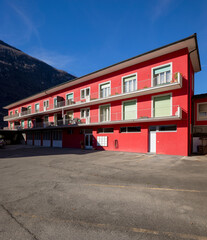 Red railing house with parking area below. Some windows open, others closed by green shutters. Blue sky and mountains behind.
