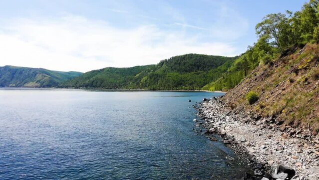Flying over the lake shore on a sunny, windless summer day. Mountains covered with forest near the shore.