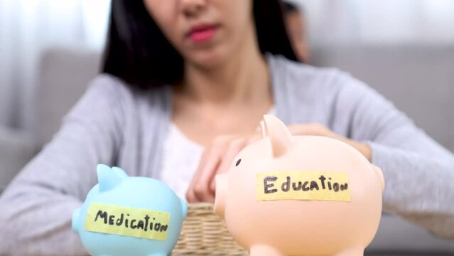 Asian worried female sitting on floor looking piggy bank represent education medication for children while kids playing behind, unsecured financial single mother tired planning for boy and girl