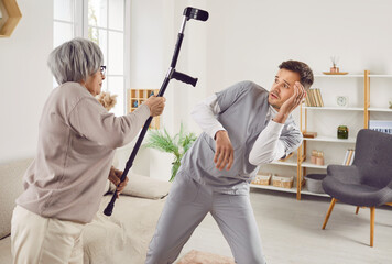 Angry elderly senior woman going to hit young male nurse or doctor with crutch. Enraged exasperated female pensioner standing at home and threatening her scared caretaker with cane. - obrazy, fototapety, plakaty