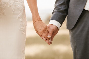 Close-up of bride and groom holding hands with wedding rings at reception outside in the backyard.