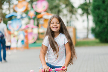 A beautiful smiling girl rides a scooter along the path of an amusement park on a summer day....