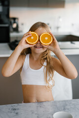 Smiling child girl covered his eyes with orange halves sitting at the table in the kitchen. Citrus...