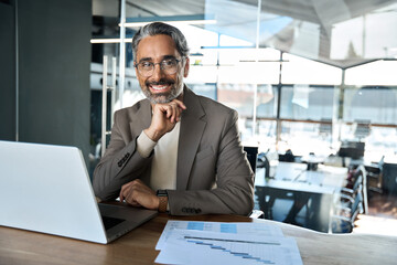 Portrait of successful mature lawyer, happy middle aged business man ceo executive, smiling older entrepreneur wearing suit and glasses looking at camera sitting at work desk with laptop in office.