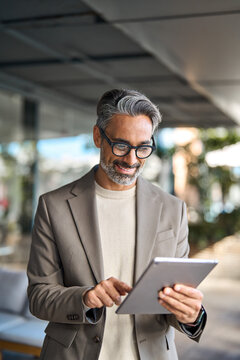 Busy happy mature older business man executive standing in office using digital tablet. Middle aged professional businessman manager wearing suit and glasses holding tab working outdoors. Vertical.