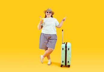 Portrait of happy smiling fat woman in hat and sunglasses with suitcase holding juice cocktail and pointing index finger to the side on yellow background. Summer holiday trip and vacation concept.