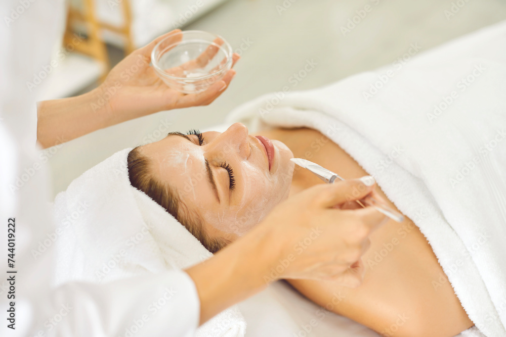 Wall mural Woman enjoying professional skin care at beauty salon. Beautician at luxury spa resort applying moisturizing or peeling mask on face of young woman in white towel. Facial treatment procedure concept