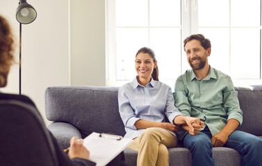 Happy smiling young couple sitting on the couch at counselor's office, listening advices and smiling holding hands. Man and woman talking with male family psychologist during therapy session.
