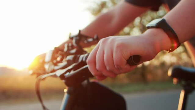 The hands of a man with a fitness bracelet hold on to the steering wheel of a bicycle against the backdrop of a summer sunset in nature. The concept of gadgets for measuring heart rate during cycling