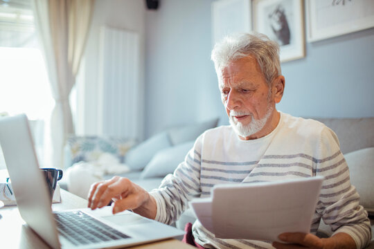 Concerned senior man using laptop and holding paperwork at home