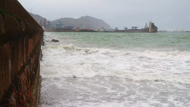 Olas rompiendo contra el muelle con el puerto industrial de fondo