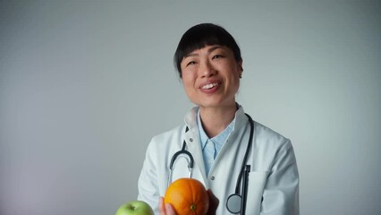 Attractive Asian female doctor with a white coat smiling and holding an apple and orange. Cheerful medical nutritionist recommending a healthy diet to a patient, 4k video footage.