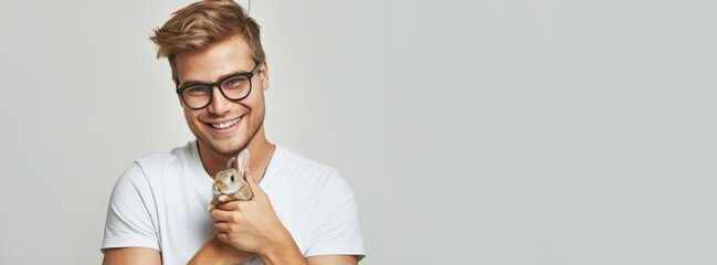 banner a smiling blond man with glasses in a white T-shirt holds an Easter bunny on a light gray background. close-up - obrazy, fototapety, plakaty