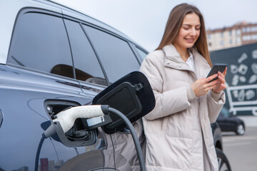 Young woman charging her electric car at the gas station, using a smartphone. Eco fuel concept. The...