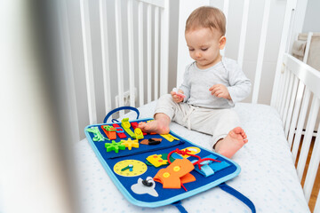 Baby playing with montessori busy book sitting in crib. Educational books and quiet books concept. Montessori busy board