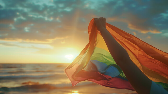 person with the big rainbow LMBTQ gay flag in his or her hand at the beach sunset