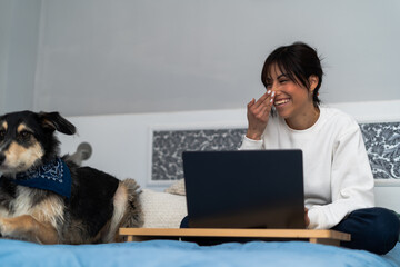 A cheerful young woman enjoys a lighthearted moment with her loyal dog while working remotely from her cozy bedroom