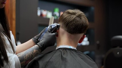 Professional barber gives stylish haircut with electric clipper, comb in modern barbershop. Tattooed hairstylist works on male client, fades sides, trims top. Grooming session, salon interior.