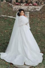 A brunette bride in a white dress with a long train holds the dress and walks down the path covered with autumn leaves. Wedding photo session in nature. Beautiful hair and makeup. Celebration