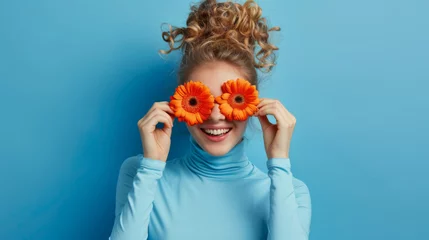 Foto op Plexiglas A person holds bright orange gerbera flowers over their eyes like glasses, smiling broadly against a blue background, creating a playful and joyful portrait. © MP Studio