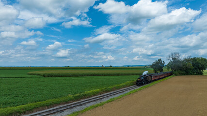 Vintage Steam Locomotive Pulling Red Passenger Cars Alongside Lush Fields And A Dirt Path Under A Broad Sky With Fluffy Clouds.