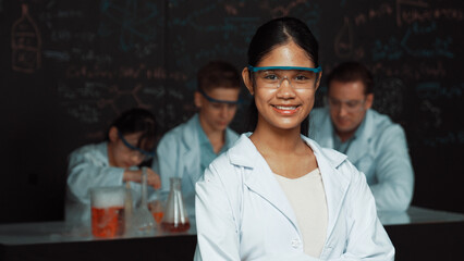 Girl looking at camera with arm folded while people doing experiment at laboratory. Cute student...