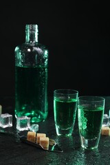 Absinthe in shot glasses, spoon, brown sugar and ice cubes on gray table against dark background....