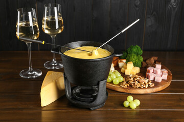 Fondue pot with melted cheese, glasses of wine and different products on wooden table