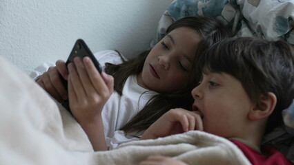 Small siblings staring at cellphone device in bed, sister sharing phone screen with brother looking at smartphone device together