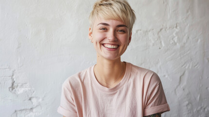 A person with a joyous smile and short hair, featuring a tattoo on their arm and piercings, is wearing a soft-colored shirt against a textured white background.