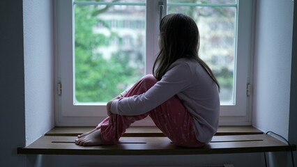 One pensive little girl seated by apartment window staring at view. 8 year old child in thoughtful...