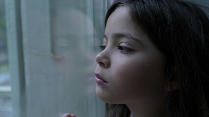Sad pensive child leaning on glass window looking outside feeling trapped at home. One little girl...
