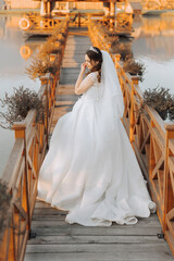 A brunette bride in a white dress with a long train holds the dress and walks on a wooden bridge. Autumn. Wedding photo session in nature. Beautiful hair and makeup. celebration