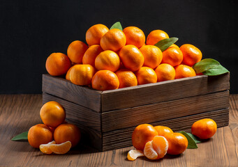 A group of orange ripe tangerines with peeled segments and green leaves lie in a wooden box on a...