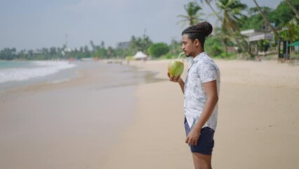 Man sips fresh coconut, strolls along tropical beach. Relaxed male enjoys palm juice on sunny coast. Casual walk, vacation vibes, sandy shore, sea wave backdrop. Solo traveler rejuvenates with coco.