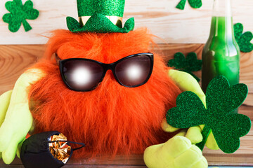 Toy leprechaun with ginger beard in green hat and sunglasses holds a shamrock in his hands. Saint...