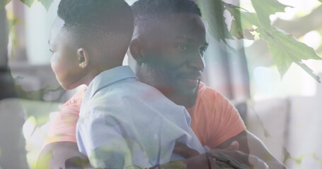 Image of leaves over happy african american father and son hugging on sofa