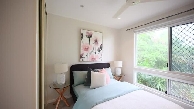 Crisp clean establishing view of single bed with ceiling fan and window overlooking tropical yard