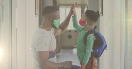 Image of lights over happy african american father and son in face masks and clapping hands